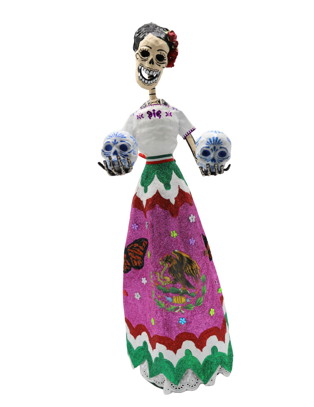A papier-mâché skeleton figurine wearing a white embroidered blouse and a vibrant pink, green, and white skirt with the Mexican coat of arms, holding two decorative skulls.