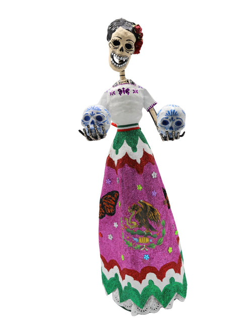A papier-mâché skeleton figurine wearing a white embroidered blouse and a vibrant pink, green, and white skirt with the Mexican coat of arms, holding two decorative skulls.