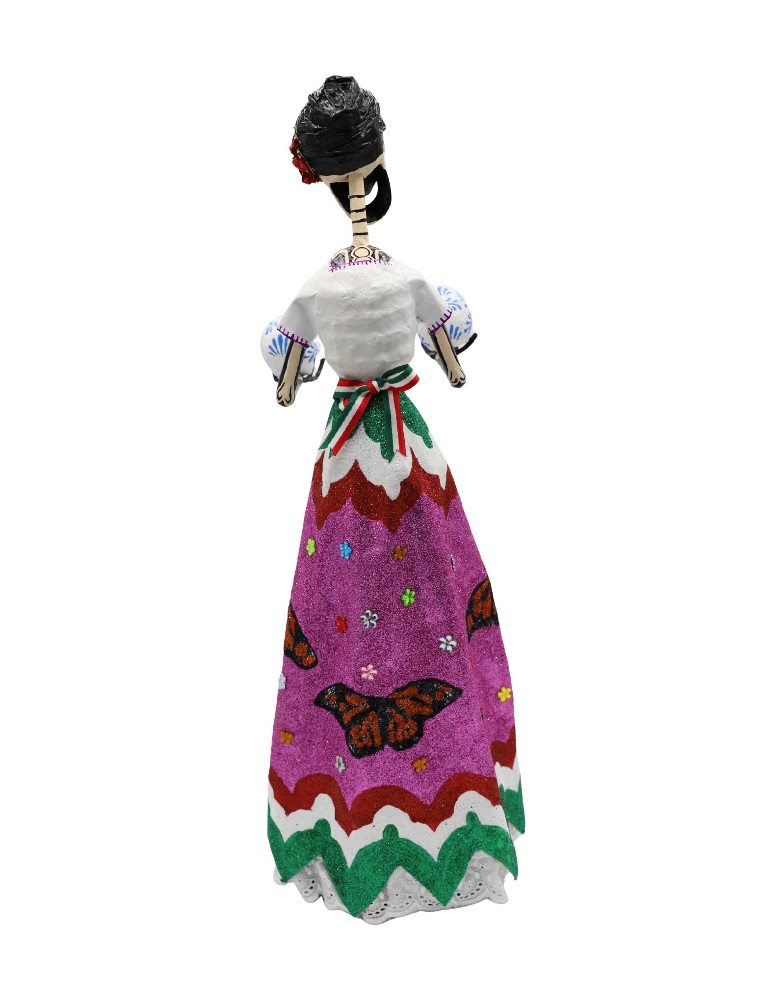 A papier-mâché skeleton figurine wearing a white embroidered blouse and a vibrant pink, green, and white skirt with the Mexican coat of arms, holding two decorative skulls.