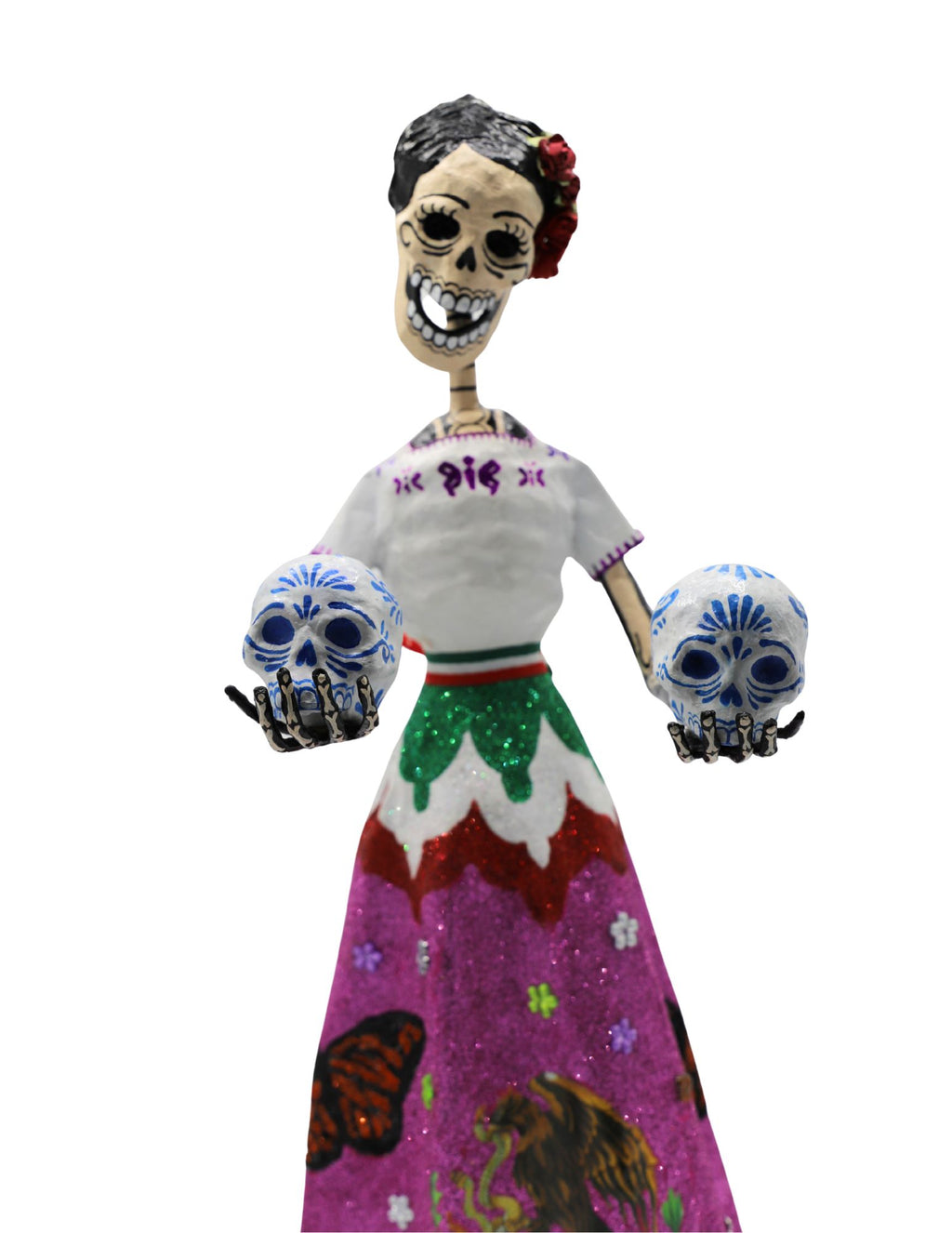 A papier-mâché skeleton figurine wearing a white embroidered blouse and a vibrant pink, green, and white skirt with the Mexican coat of arms, holding two decorative skulls.