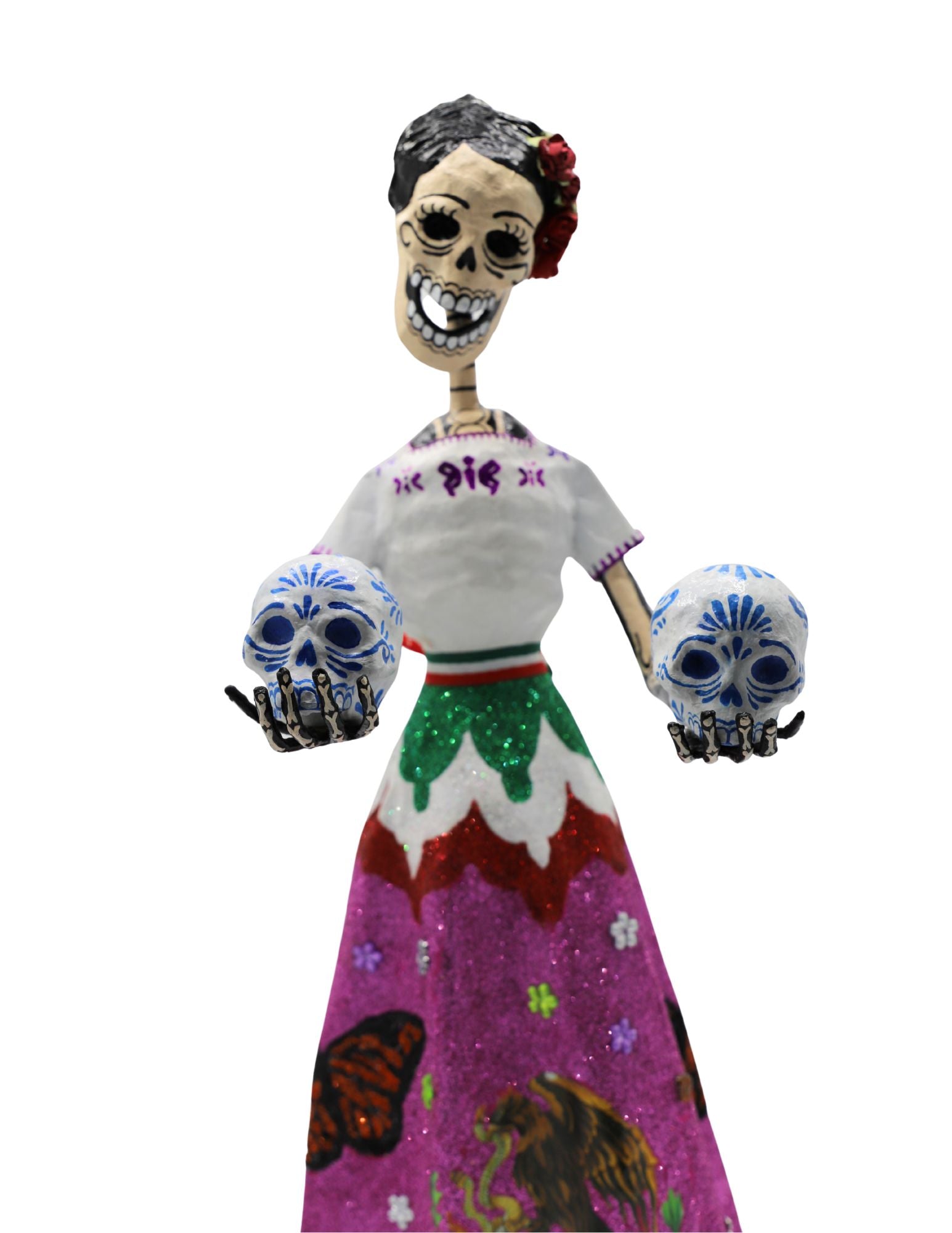 A papier-mâché skeleton figurine wearing a white embroidered blouse and a vibrant pink, green, and white skirt with the Mexican coat of arms, holding two decorative skulls.