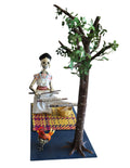 A papier-mâché sculpture featuring a skeleton woman weaving a rug next to a tree, with a small rooster at the base, all mounted on a dark blue rectangular platform.