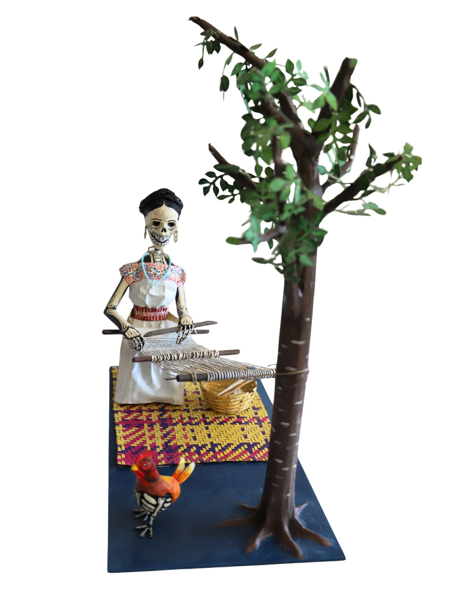 A papier-mâché sculpture featuring a skeleton woman weaving a rug next to a tree, with a small rooster at the base, all mounted on a dark blue rectangular platform.