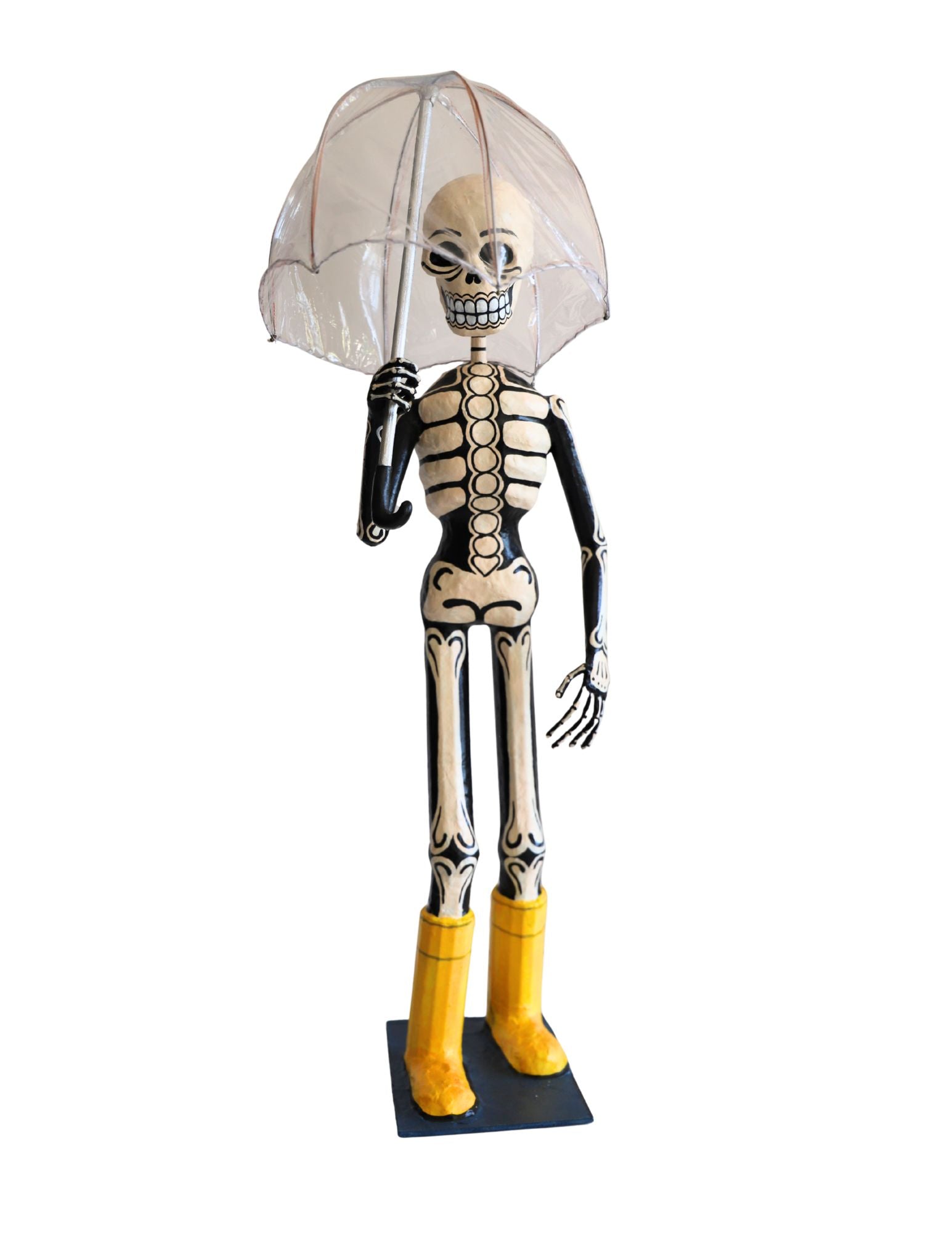 A papier-mâché skeleton figurine holding a clear transparent umbrella, standing on a black base. The skeleton has black and white painted bones with bright yellow boots.