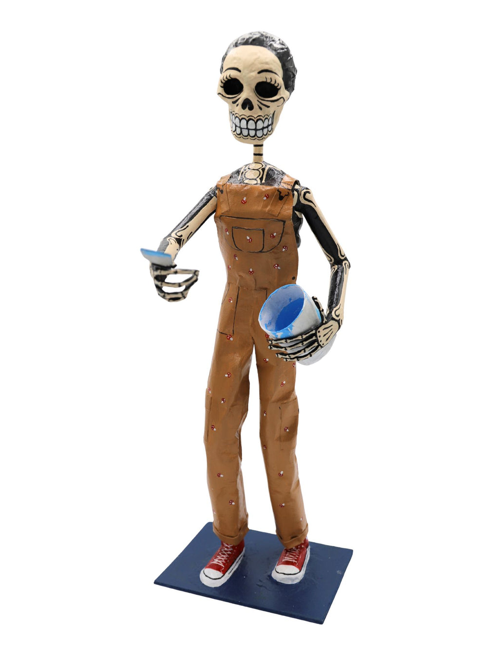 A papier-mâché skeleton figurine dressed in brown overalls and red sneakers, holding a blue cup.