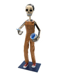 A papier-mâché skeleton figurine dressed in brown overalls and red sneakers, holding a blue cup.