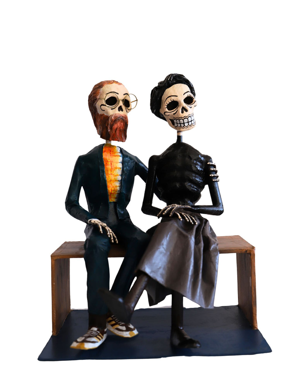 A papier-mâché figurine of a skeleton couple sitting on a wooden bench. The man has a reddish-brown beard and mustache, wearing a dark suit with a bright orange shirt. The woman has black hair, a black top, and grey skirt. 