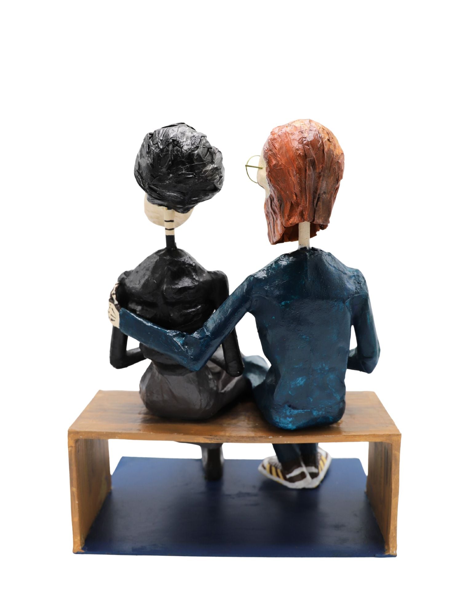 A papier-mâché figurine of a skeleton couple sitting on a wooden bench. The man has a reddish-brown beard and mustache, wearing a dark suit with a bright orange shirt. The woman has black hair, a black top, and grey skirt. 