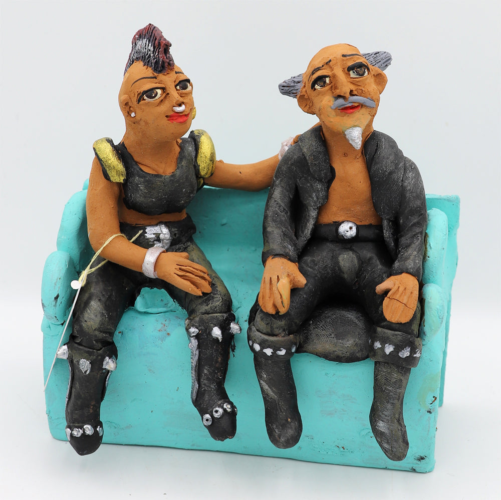 A ceramic sculpture of two figurines of a man and a woman wearing edgy clothing sitting on a blue couch.
