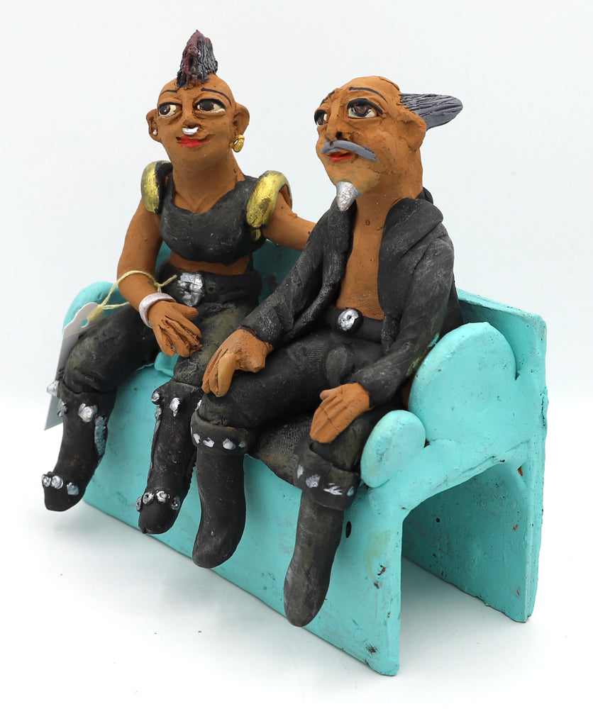A ceramic sculpture of two figurines of a man and a woman wearing edgy clothing sitting on a blue couch.