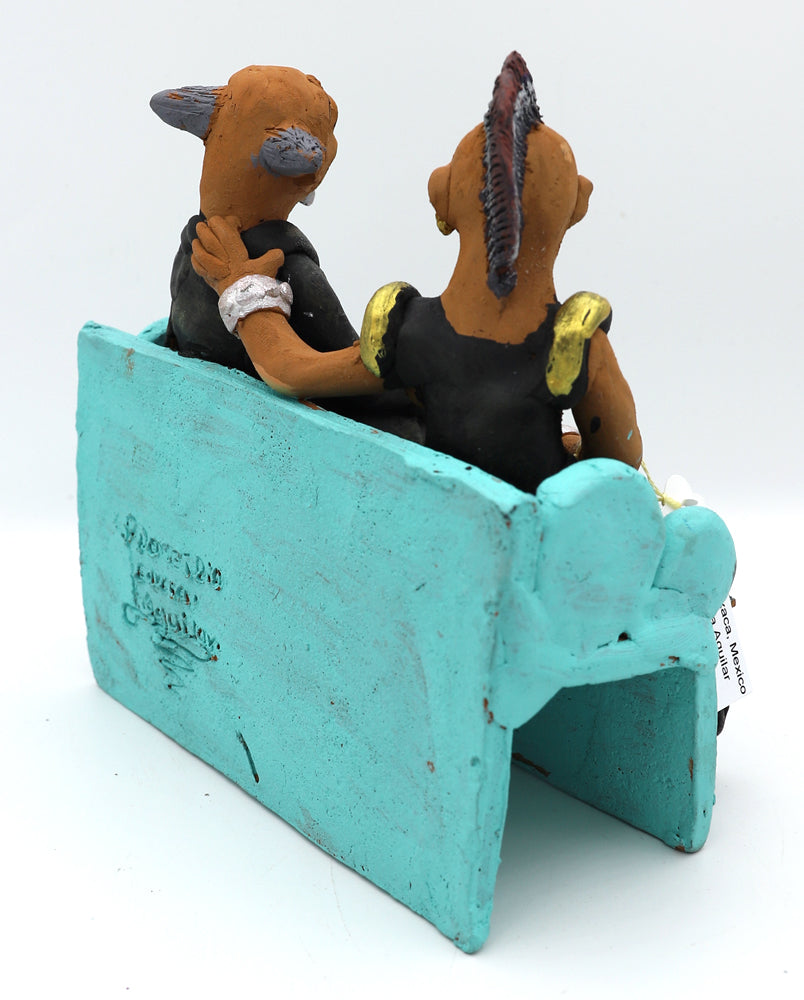 A ceramic sculpture of two figurines of a man and a woman wearing edgy clothing sitting on a blue couch.