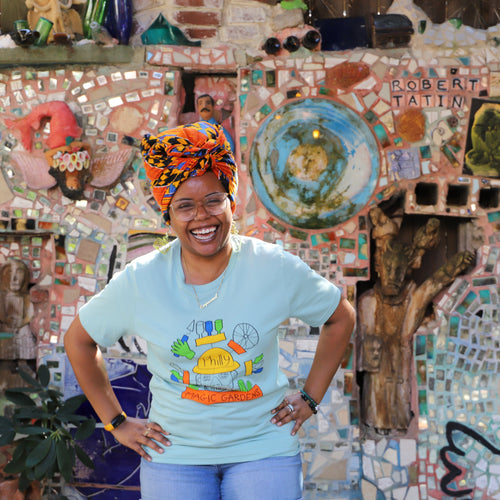 A woman laughs wearing a t-shirt depicting a colorful drawing of Isaiah Zagar's head with bike wheels and bottles surrounding it. 