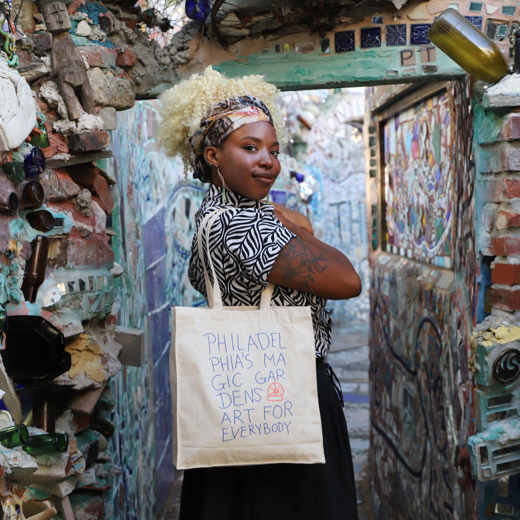 A woman models a beige tote bag with the words "Philadelphia's Magic Gardens, Art For Everybody" on it.