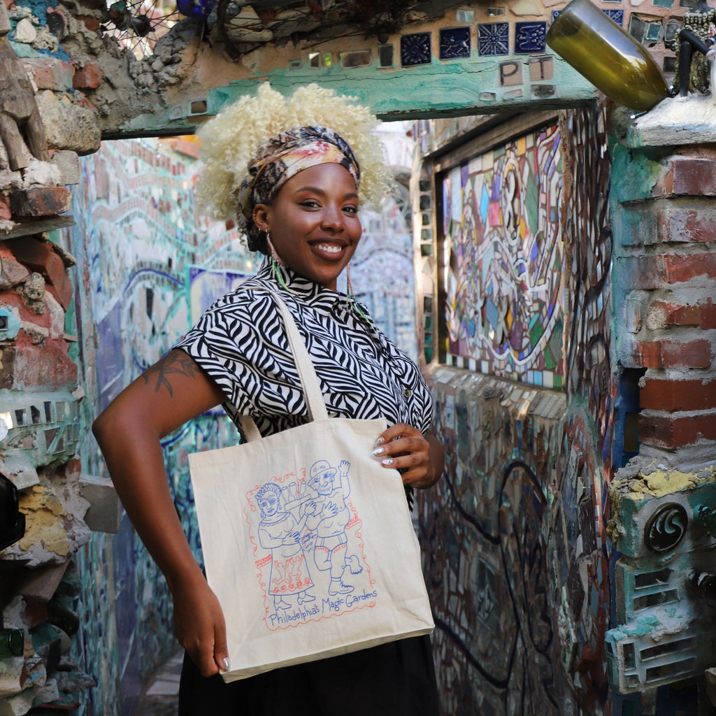 A woman smiles while modeling a beige tote bag with a drawing of Isaiah and Julia Zagar on it.