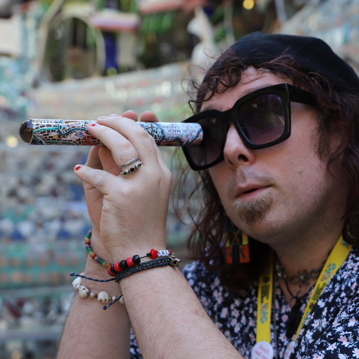 A person with long curly hair, a black beret, and black sunglasses looks through a decorative, patterned metal kaleidoscope. They are wearing a black and white floral shirt, a yellow lanyard, and multiple bracelets, set against a highly detailed, colorful mosaic background.