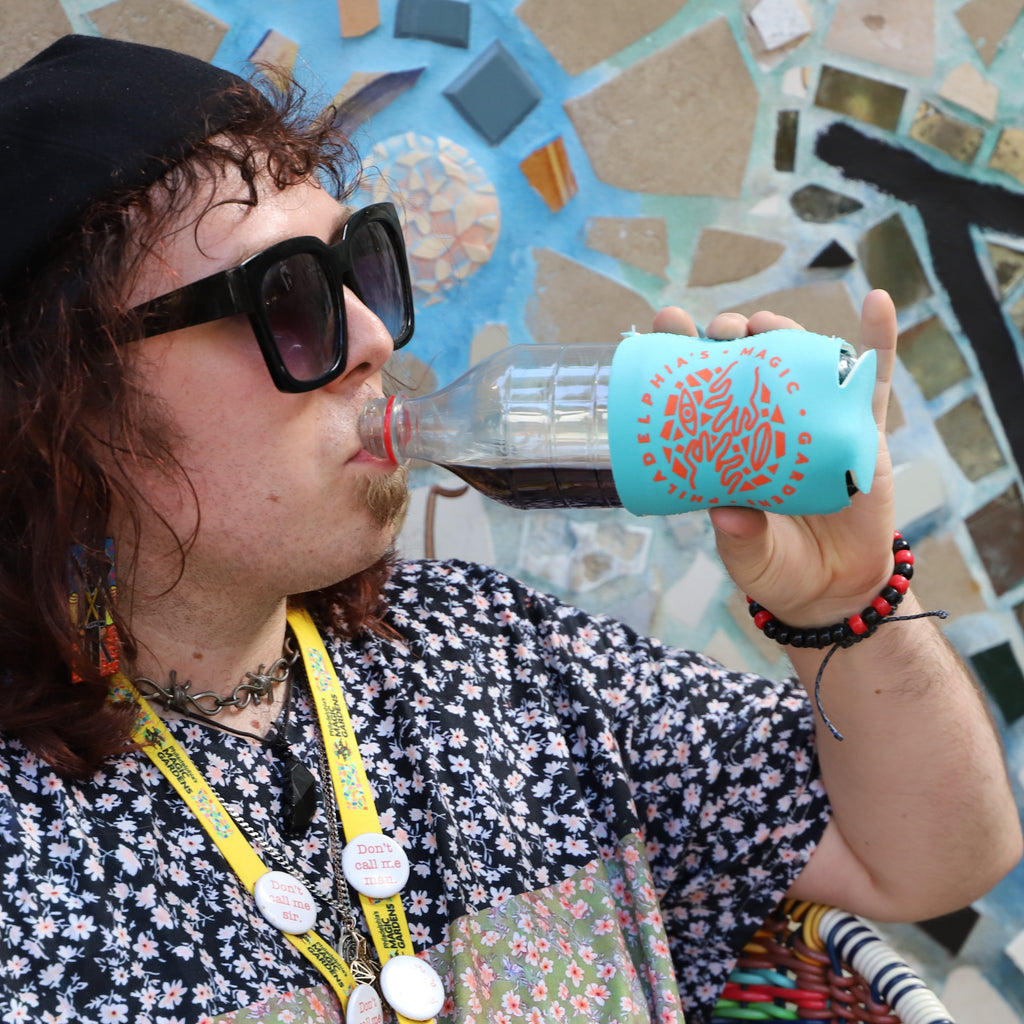 A woman in sunglasses, a black beret, and a patterned shirt drinks out of a bottle using a teal koozie with Philadelphia's Magic Gardens' logo on it.