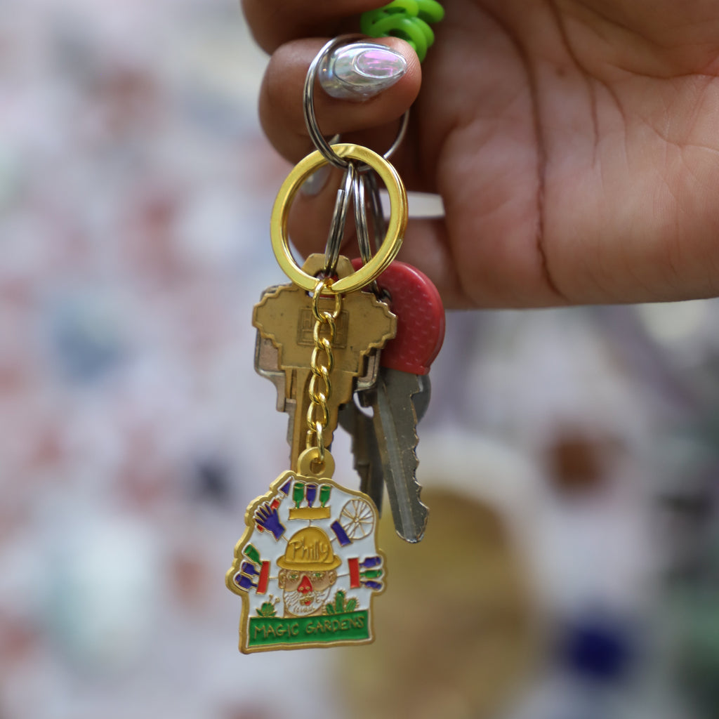 A close-up of a hand holding a gold-colored keyring with a small colorful religious charm and several keys, set against a heavily blurred, patterned background.