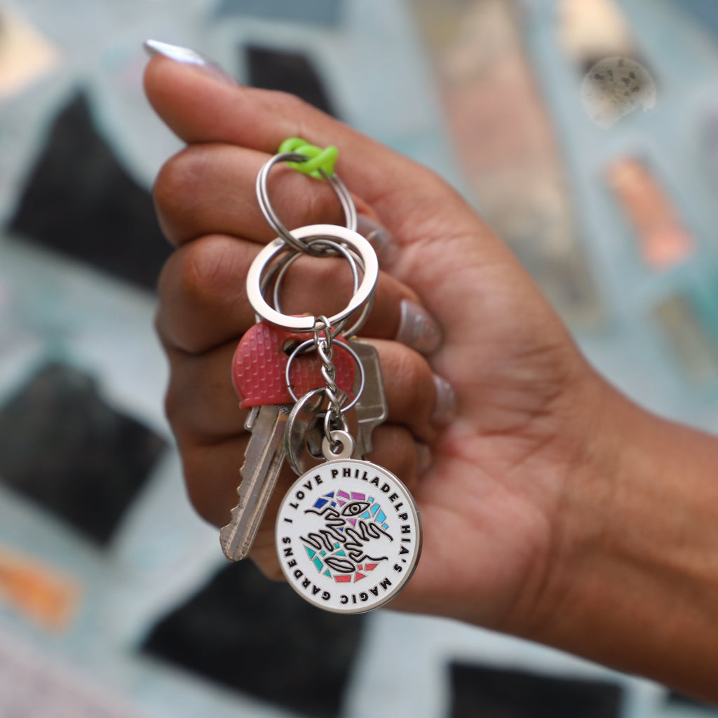 A hand holds a key ring with a round keychain that reads "I love Philadelphia's Magic Gardens" on it. 