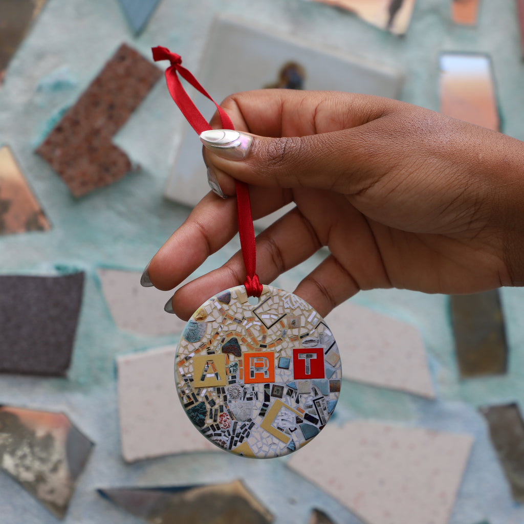 A hand holds a circular ornament with a mosaicked design on the front and a red ribbon. 