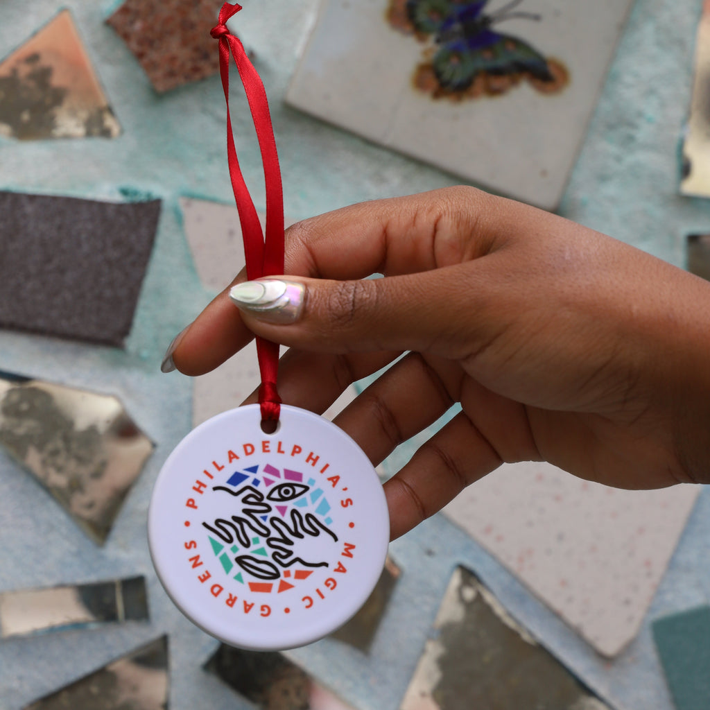 A hand holds a circular ornament with Philadelphia's Magic Gardens logo on it and a red ribbon. 
