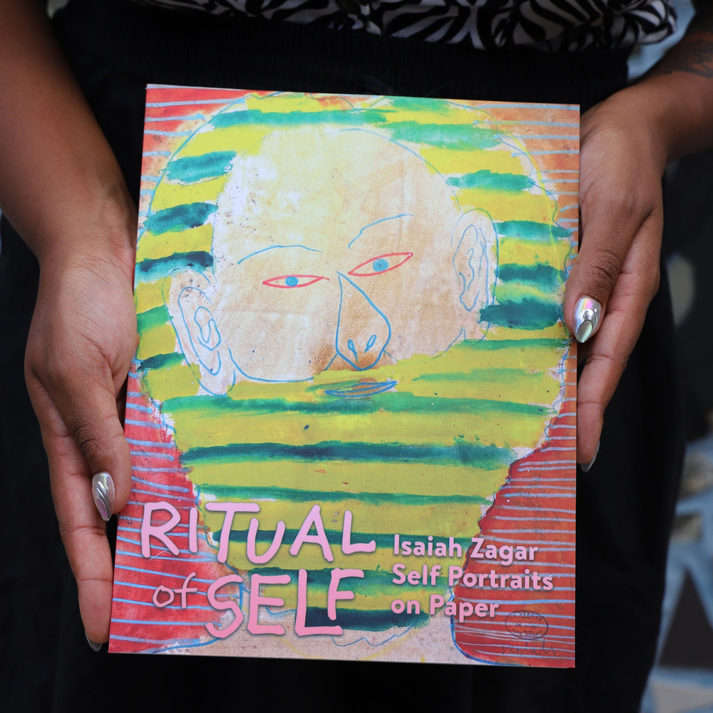 Hands hold a book with a self-portrait of artist Isaiah Zagar on the front. The title of the book is 'Ritual of Self: Isaiah Zagar Self Portraits on Paper.'