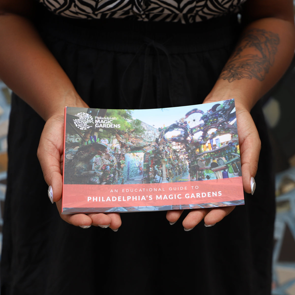 A person holding a book titled 'An Educational Guide to Philadelphia's Magic Gardens' in front of a colorful mosaic tile wall.