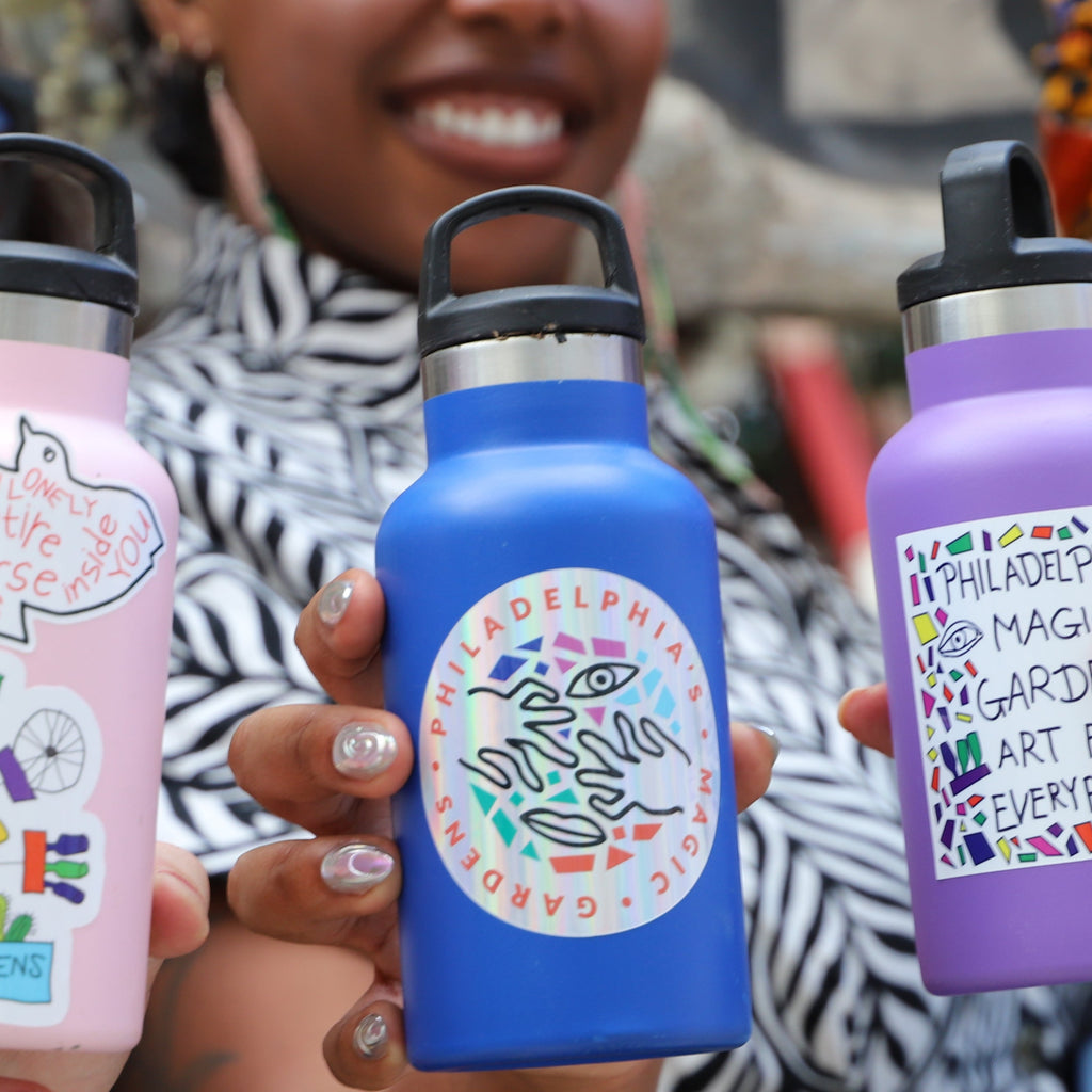 A blue water bottle with a round sticker on it containing the logo for Philadelphia's Magic Gardens