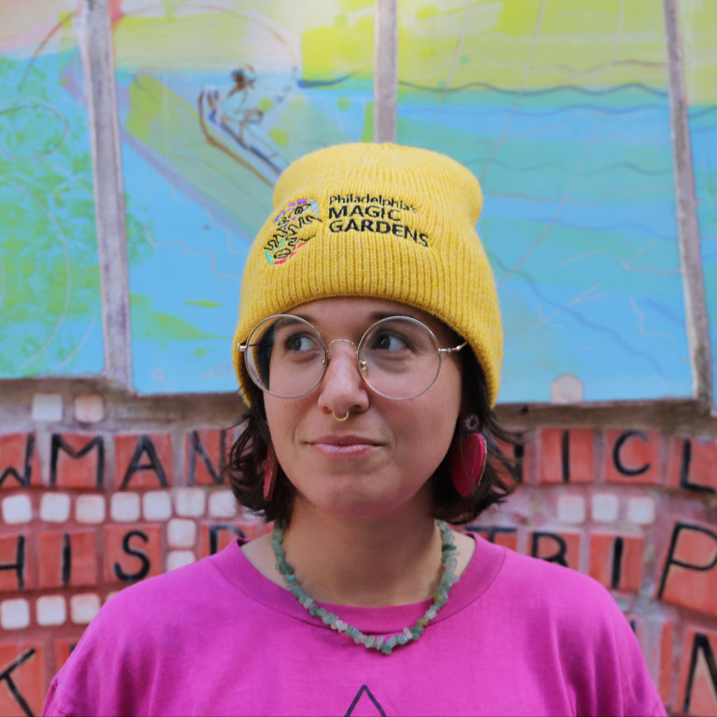 A woman in a pink shirt and glasses wearing a yellow beanie with the logo for Philadelphia's Magic Gardens on it. 