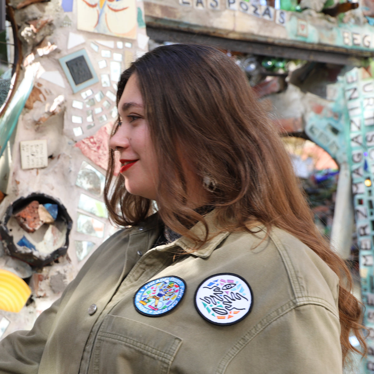 A woman with long hair wearing a jacket with two Philadelphia's Magic Gardens patches on it in front of a mosaicked background. 
