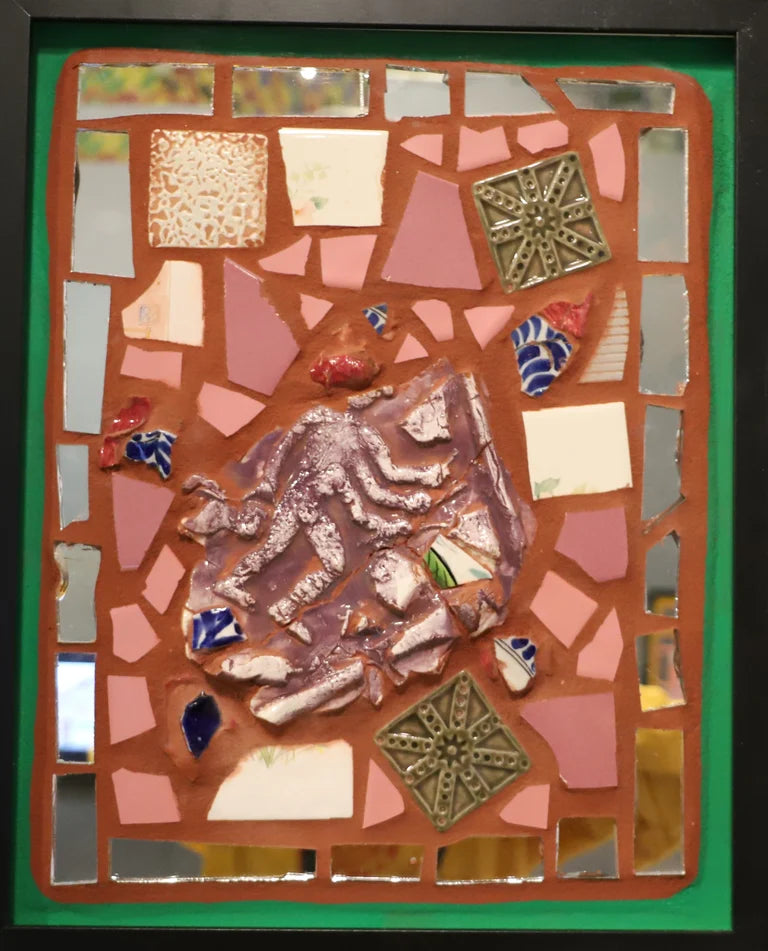Framed mosaic with various tiles and mirrors on orange grout with a green border.