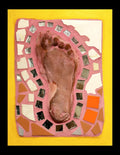 A small mosaic featuring the mold of a foot surrounded by mirror pieces and tiles on pink grout with a yellow border.